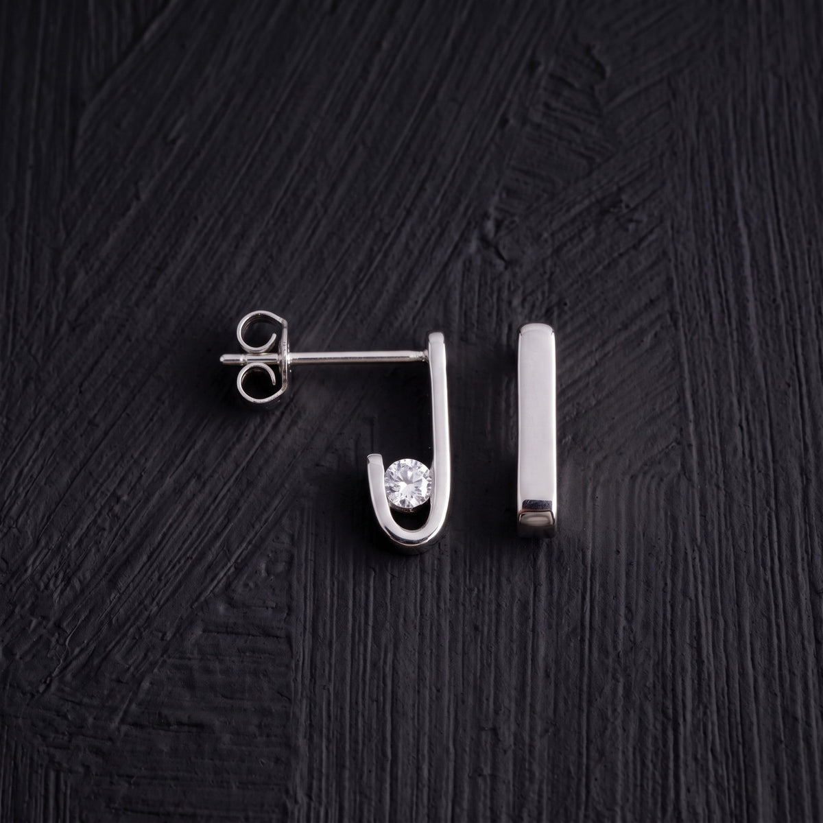 Pair of J-02 earrings on a black background: one from the side with its zircon-set curve, the other from the front as a straight line