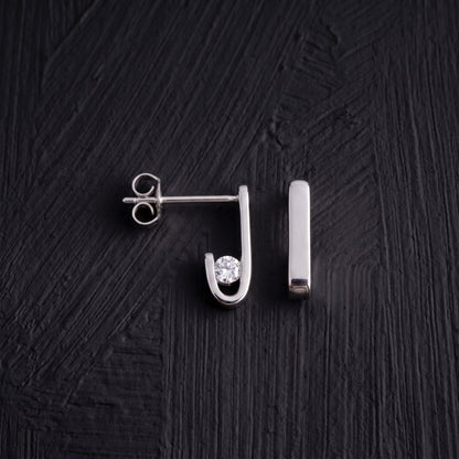 Pair of J-02 earrings on a black background: one from the side with its zircon-set curve, the other from the front as a straight line