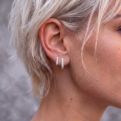 Two J earrings worn side by side on the ear of a woman with short blonde hair: the long J-02 and a shorter J earring in a second piercing, seen from the front as two straight silver lines