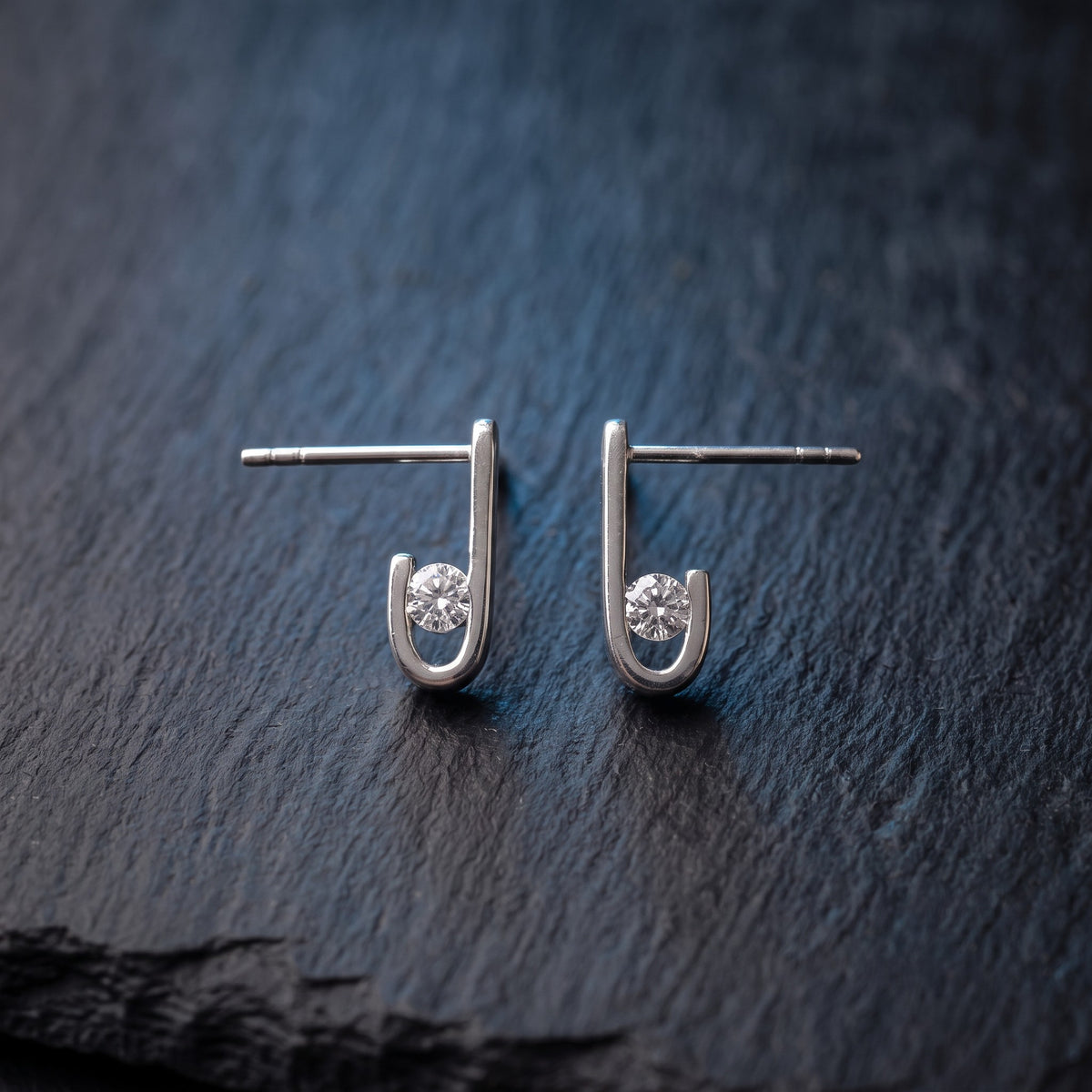 Pair of J-02 earrings on dark stone, side view: clear zircons seated in the J curve, with the earring posts