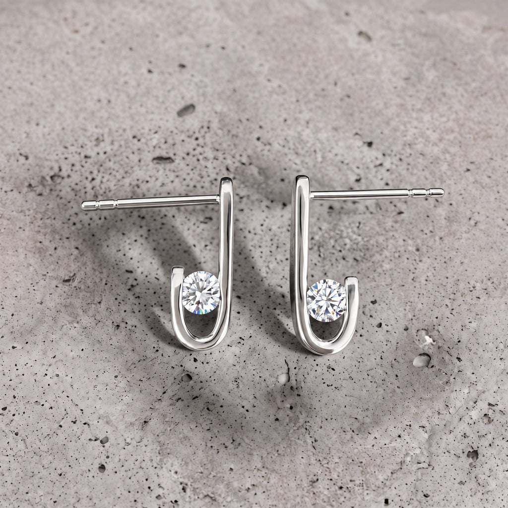 Close-up of a pair of J-02 earrings on concrete: a round clear zircon inside the J curve