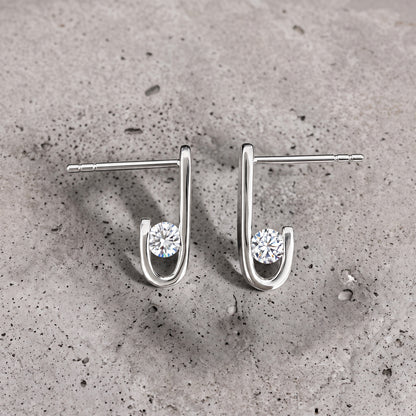 Close-up of a pair of J-02 earrings on concrete: a round clear zircon inside the J curve
