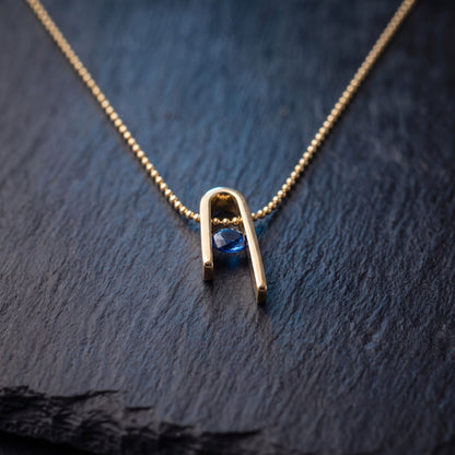 Close-up of the Minimalist n Necklace on dark stone: the n form, the blue zircon and the bead chain