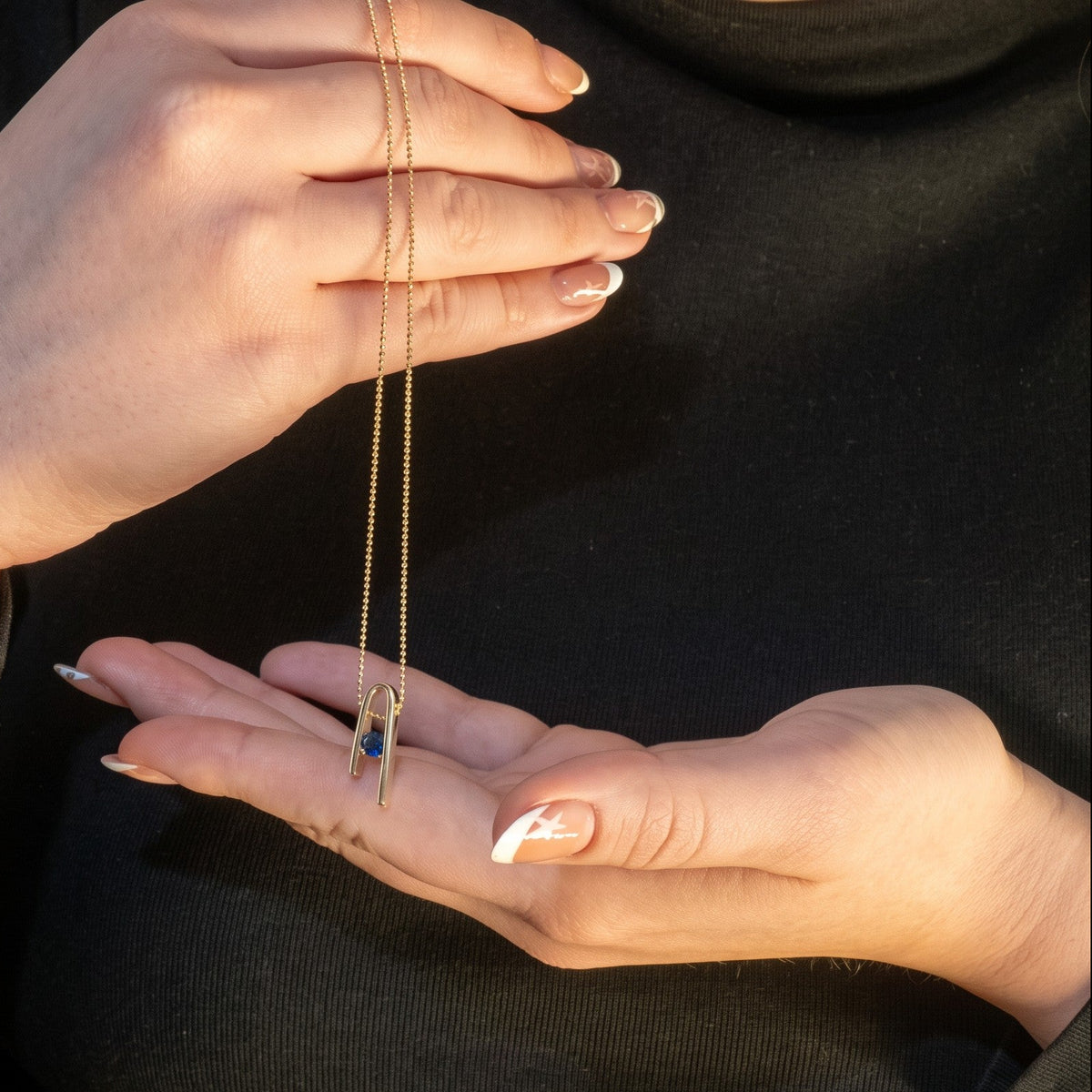 14K gold plated Minimalist n Necklace hanging between two hands, against a black sweater